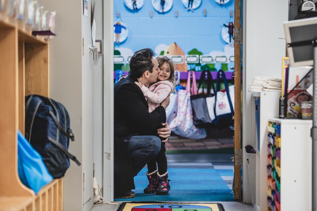 A parent kneels in a classroom doorway to kiss a small girl in a pink coat goodbye