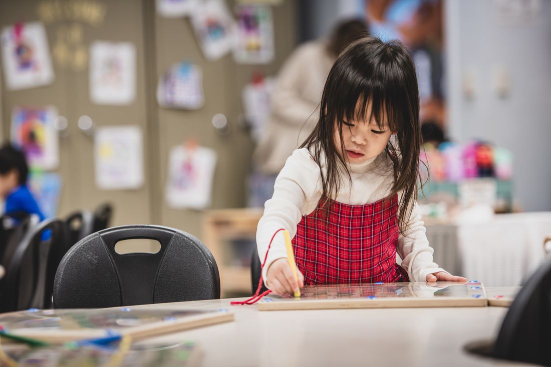 A girl in a red plaid pinafore holds a pencil and concentrates on a puzzle board