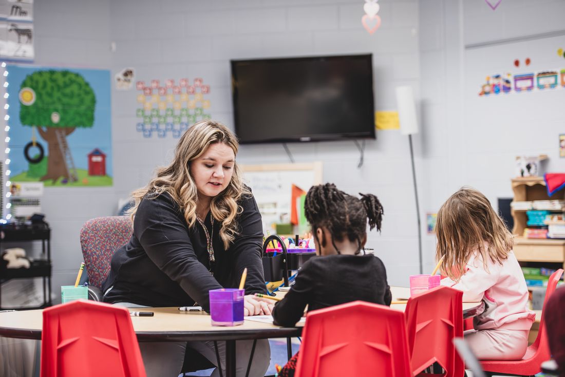 A teacher at a low table holds up a picture for children sitting in red chairs