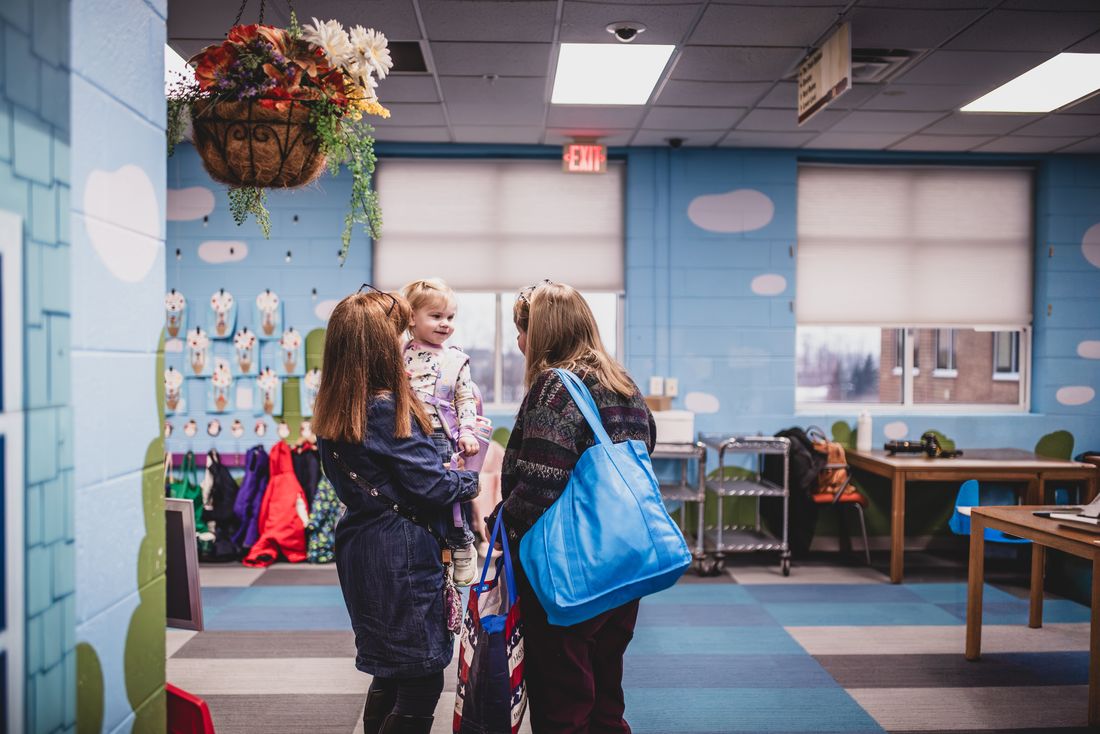 A mother holds her toddler while chatting with a teacher in a bright classroom with sky-blue cloud walls