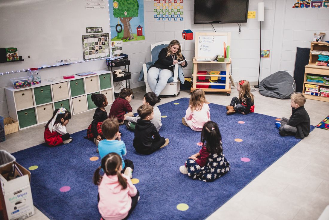 Children sit in a circle on a blue rug while a teacher leads from an armchair