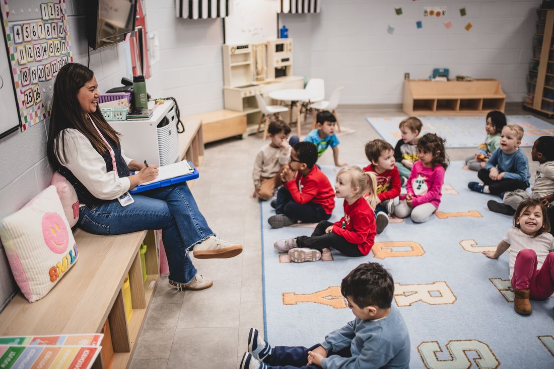 A teacher with a clipboard sits on a bench while her class sits on an alphabet rug, one girl grinning at the camera